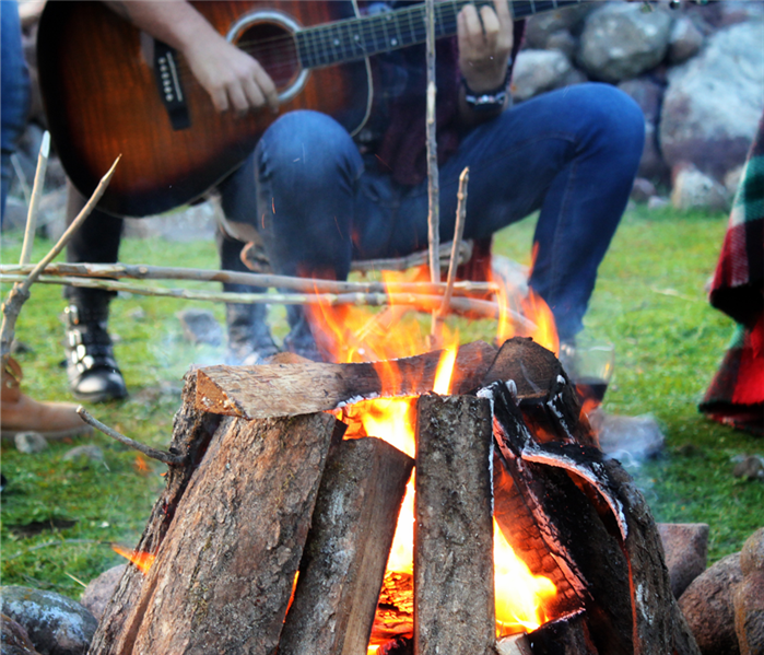 A man playing a guitar sits behind a campfire.