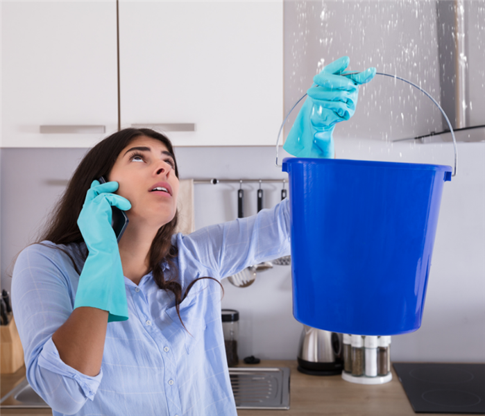 Picture shows a woman holding a blue bucket up to the celing catching droplets of water leaking. 